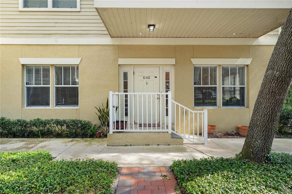 A ground-level apartment entrance features a white door, a small porch, and surrounding landscaping — 9177 SW 52ND ROAD UNIT E-103, GAINESVILLE, FL