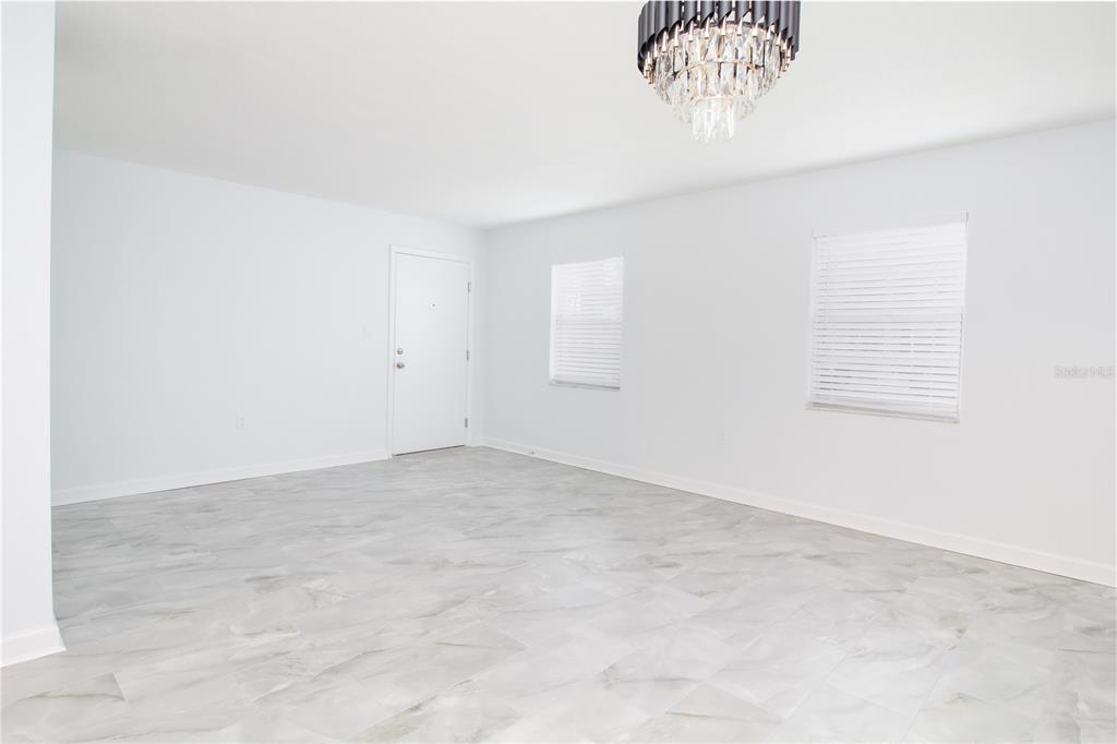 An empty room features white walls, light gray marble-look tile flooring, and a crystal chandelier — 1115 RUE DES CHATEAUX UNIT 1A, SOUTH PASADENA, FL