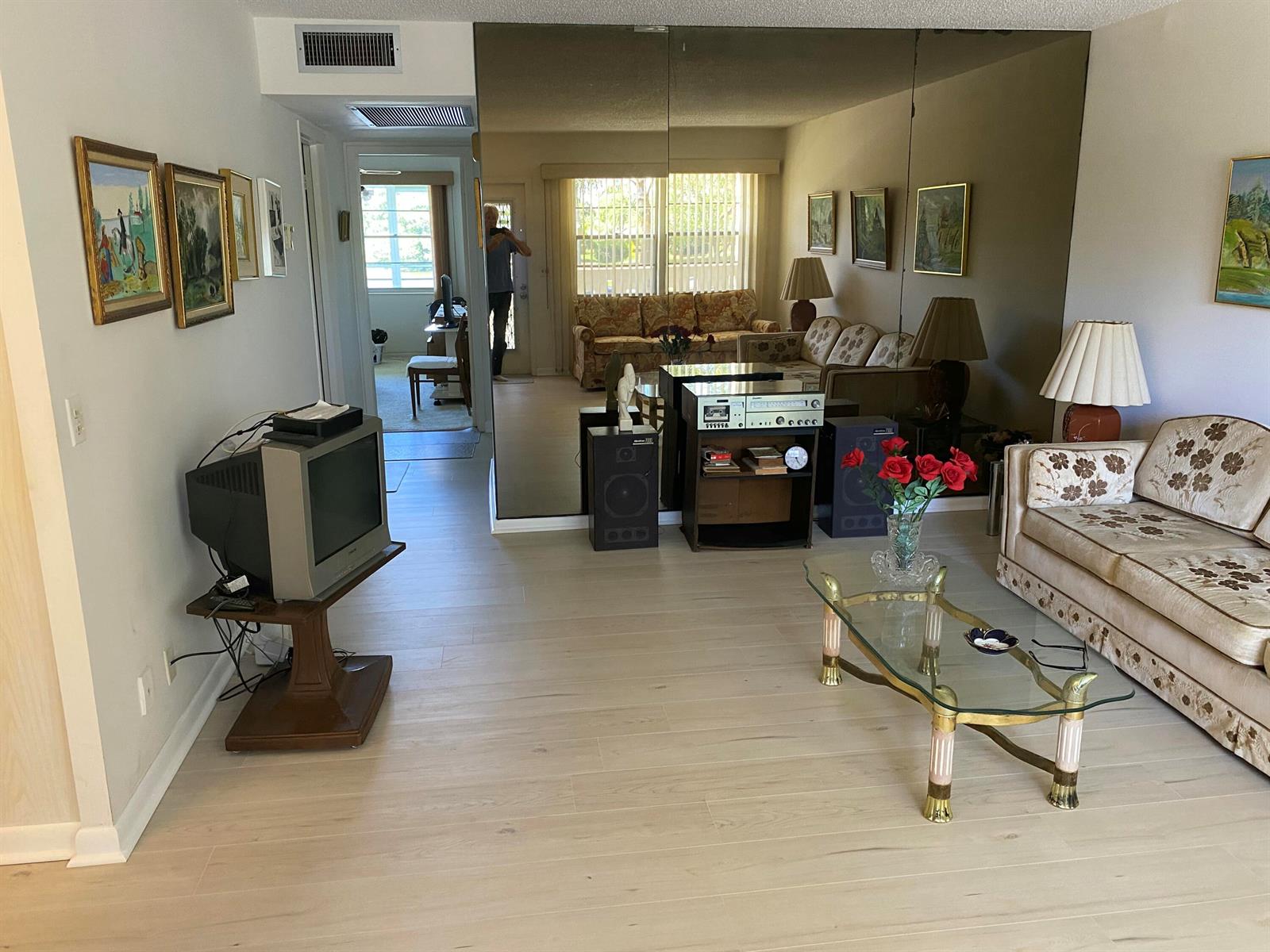 A living room features light wood flooring, a mirrored wall, a sofa, and a television — 2054 Ainslie D, Boca Raton FL 33434, BOCA RATON, FL