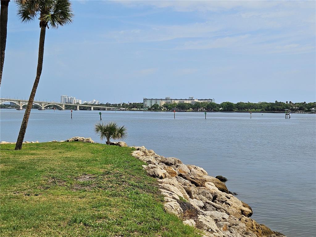 A grassy shoreline with rocks overlooks a wide river, a bridge, and distant buildings — 632 MARINA POINT DRIVE UNIT 6320, DAYTONA BEACH, FL