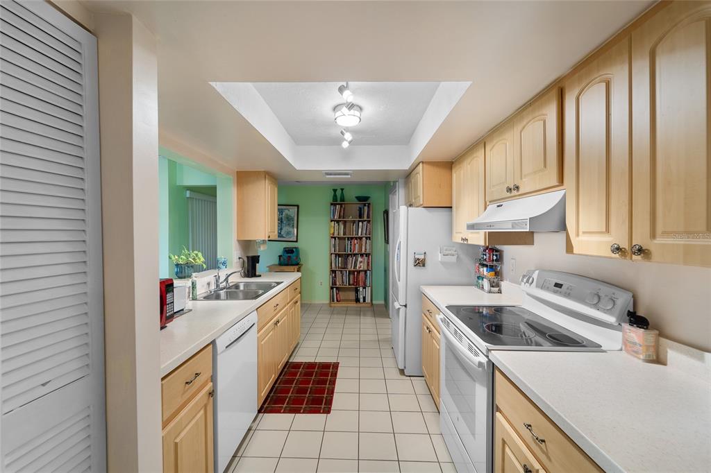 A galley kitchen features light wood cabinets, white countertops, white appliances, and a tiled floor — 6712 11TH AVENUE W UNIT 5601, BRADENTON, FL