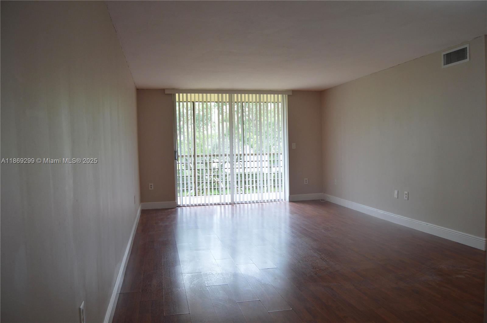 An empty room features dark wood-look flooring, beige walls, and a sliding glass door — 3490 Foxcroft Rd # B212, Miramar FL 33025, MIRAMAR, FL
