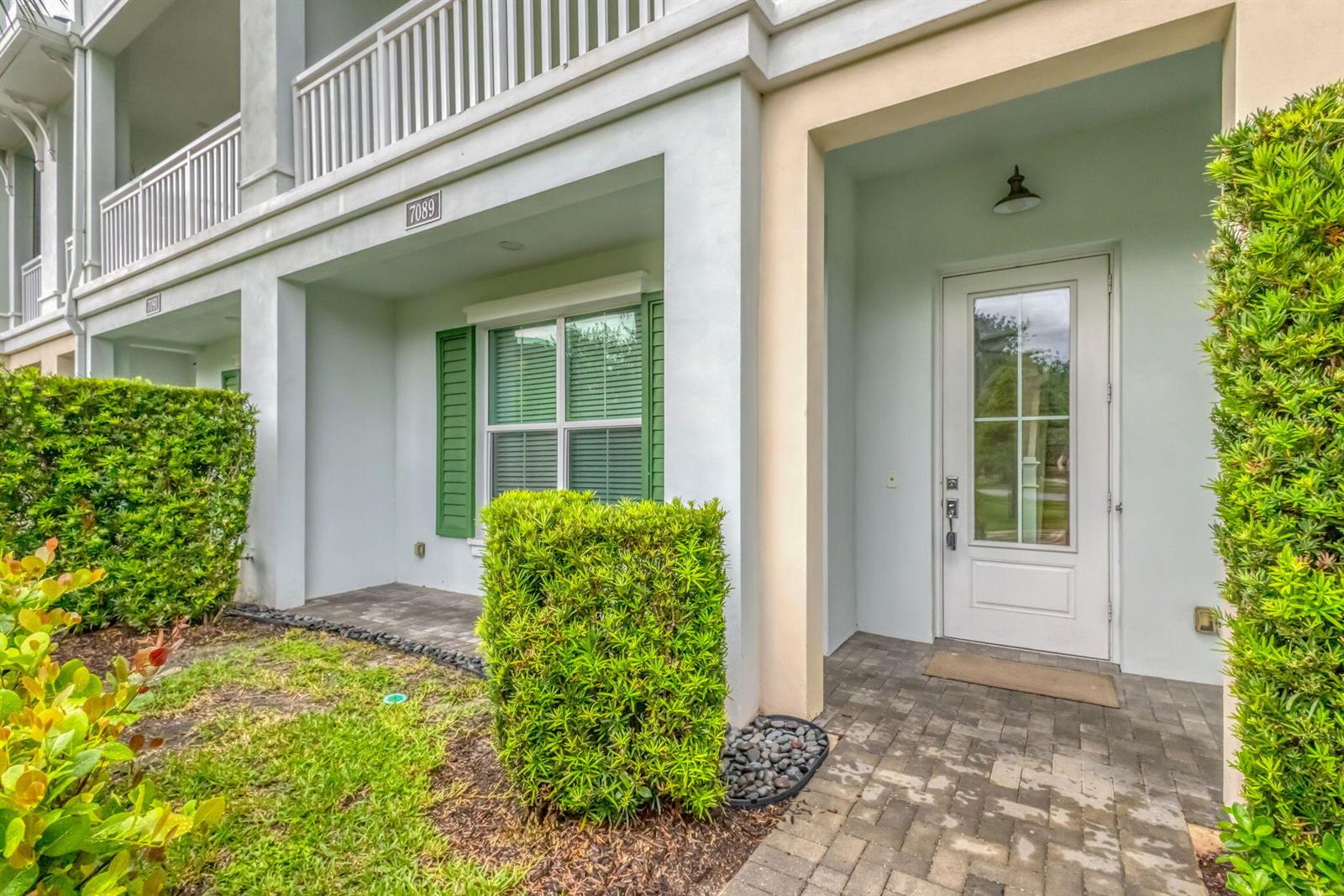 A white building exterior features a front door, a window with green shutters, and landscaping — 7089 Edison Place, Palm Beach Gardens FL 33418, PALM BEACH GARDENS, FL