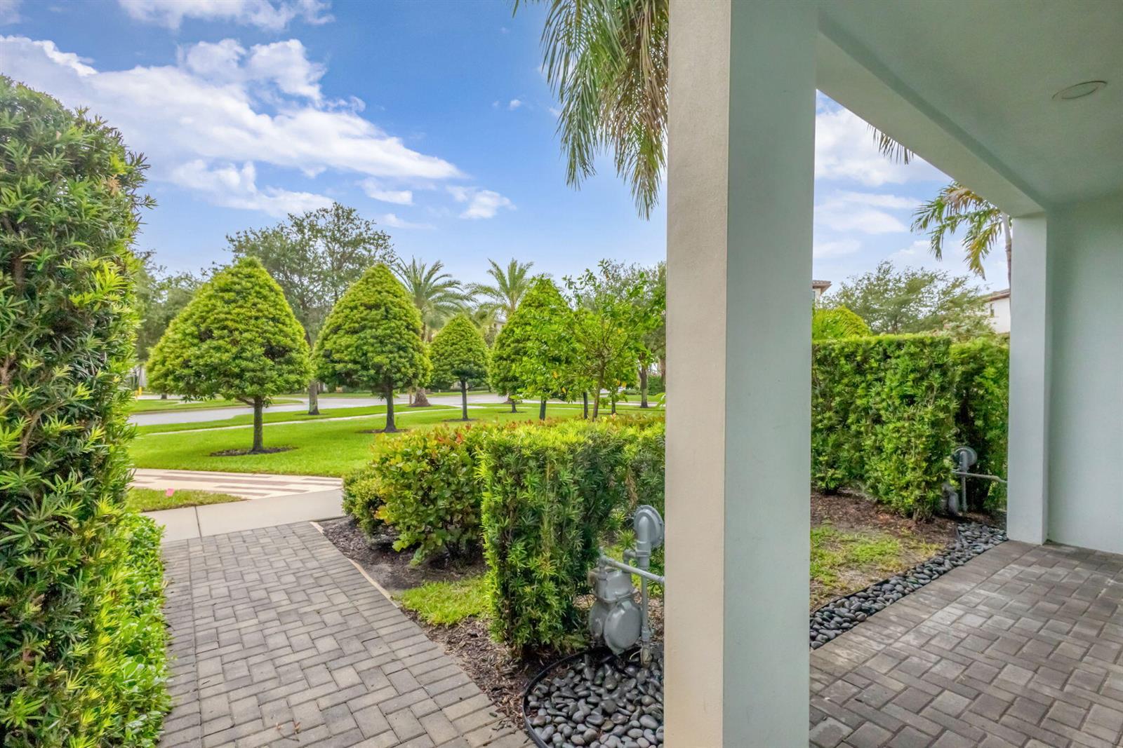A paved walkway leads from a covered porch past manicured hedges toward a grassy neighborhood — 7089 Edison Place, Palm Beach Gardens FL 33418, PALM BEACH GARDENS, FL