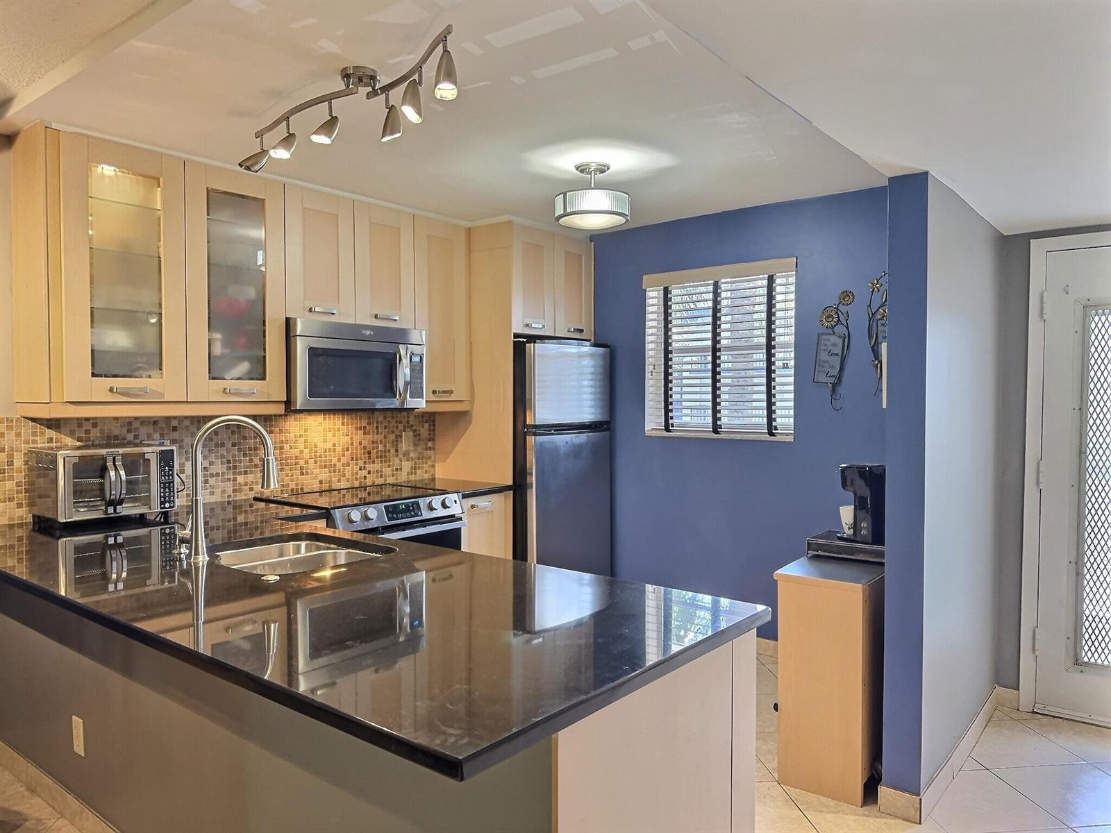 A kitchen features light wood cabinets, black countertops, a blue accent wall, and stainless appliances — 3150 Holiday Springs Boulevard # 204, Margate FL 33063, MARGATE, FL