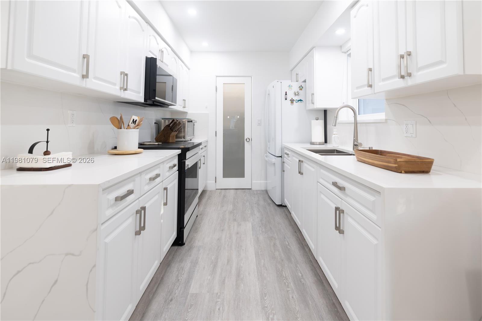 A galley kitchen features white cabinets, quartz countertops, stainless steel appliances, and light wood flooring — 19600 Gulfstream Rd, Cutler Bay FL 33157, CUTLER BAY, FL