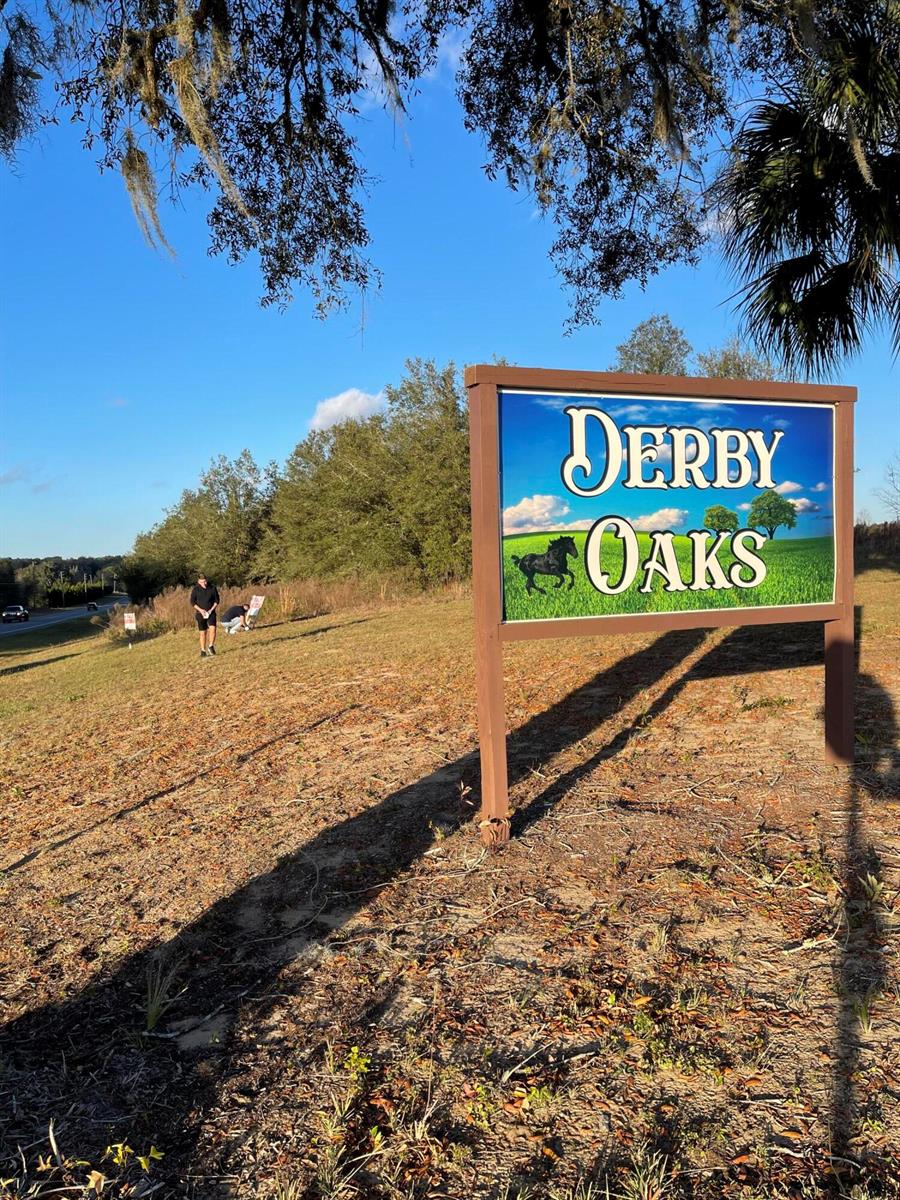 A wooden sign for Derby Oaks stands on a grassy hill with two people nearby — 7451 E Derby Oaks Drive, FL 34436, FL