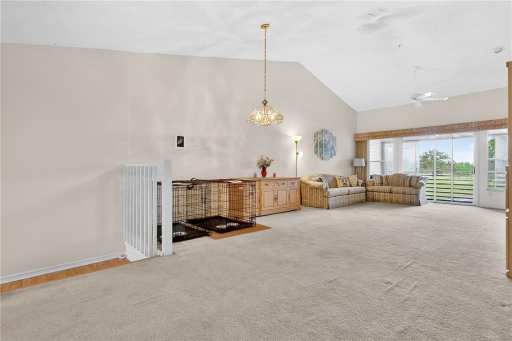 A spacious living room with vaulted ceilings, beige carpeting, a dog crate, and sliding glass doors — 6126 SUNNYVALE DRIVE UNIT 2303, ORLANDO, FL