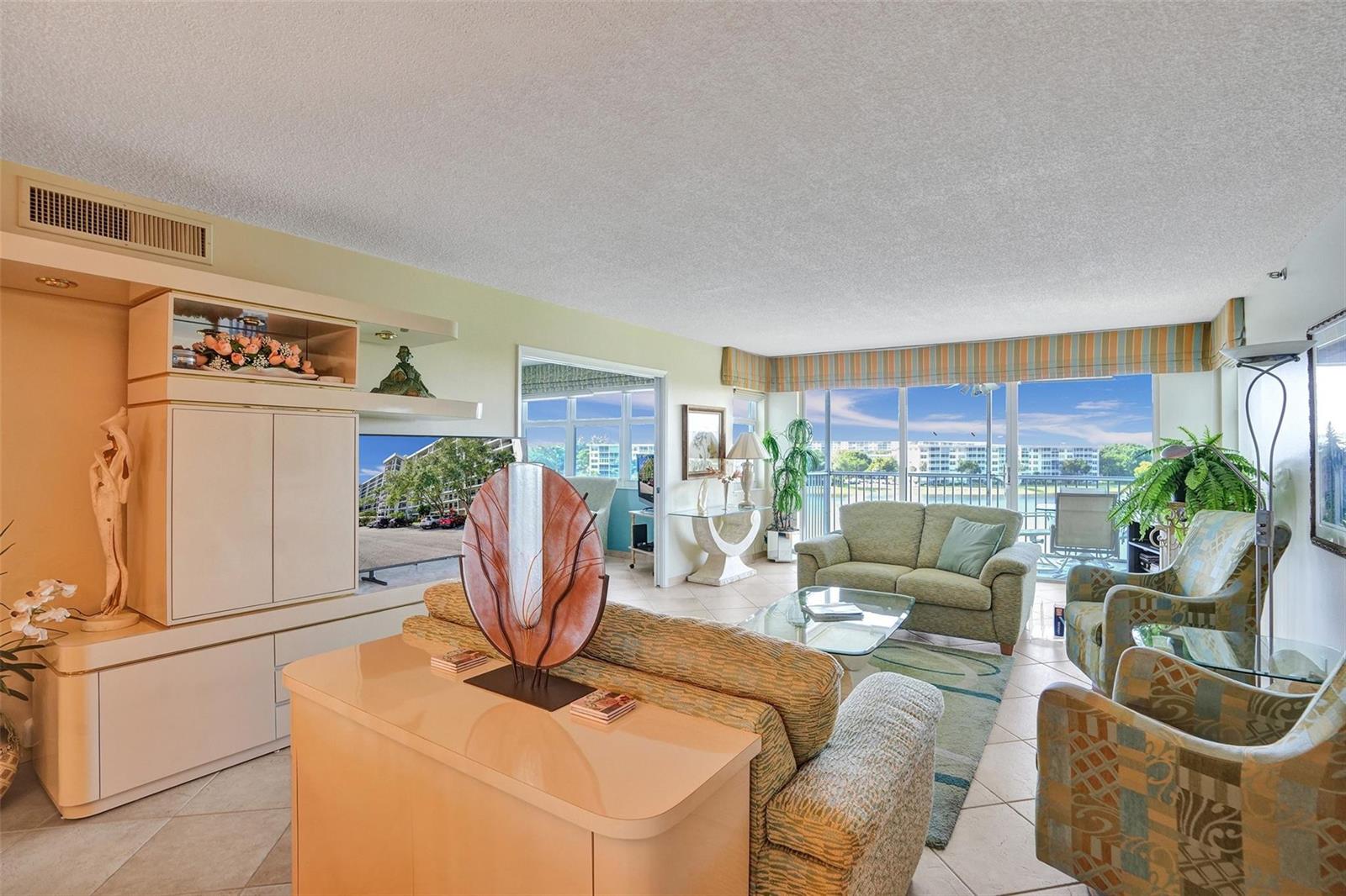 A living room features a large entertainment center, patterned furniture, and a balcony with water views — 2900 N Course Drive # 602, Pompano Beach FL 33069, POMPANO BEACH, FL