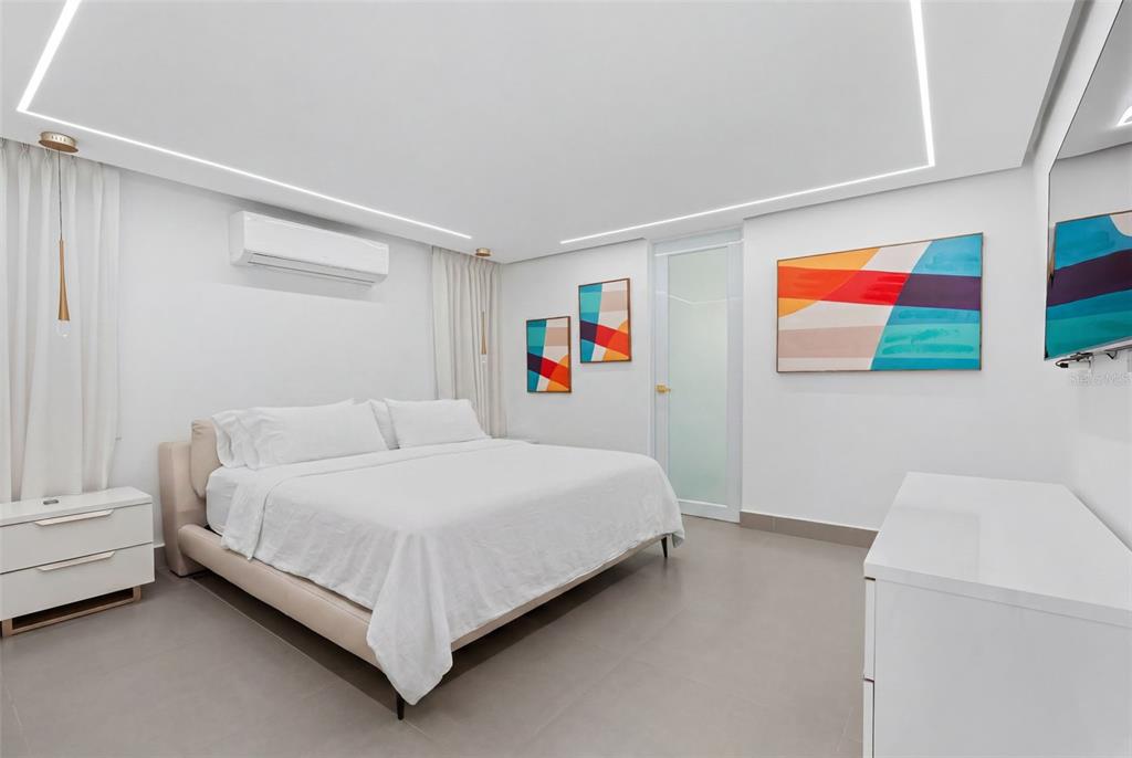 A modern bedroom features a white bed, minimalist furniture, abstract wall art, and recessed lighting. k — 96 CALLE AMAZONAS, BAYAMON, PR