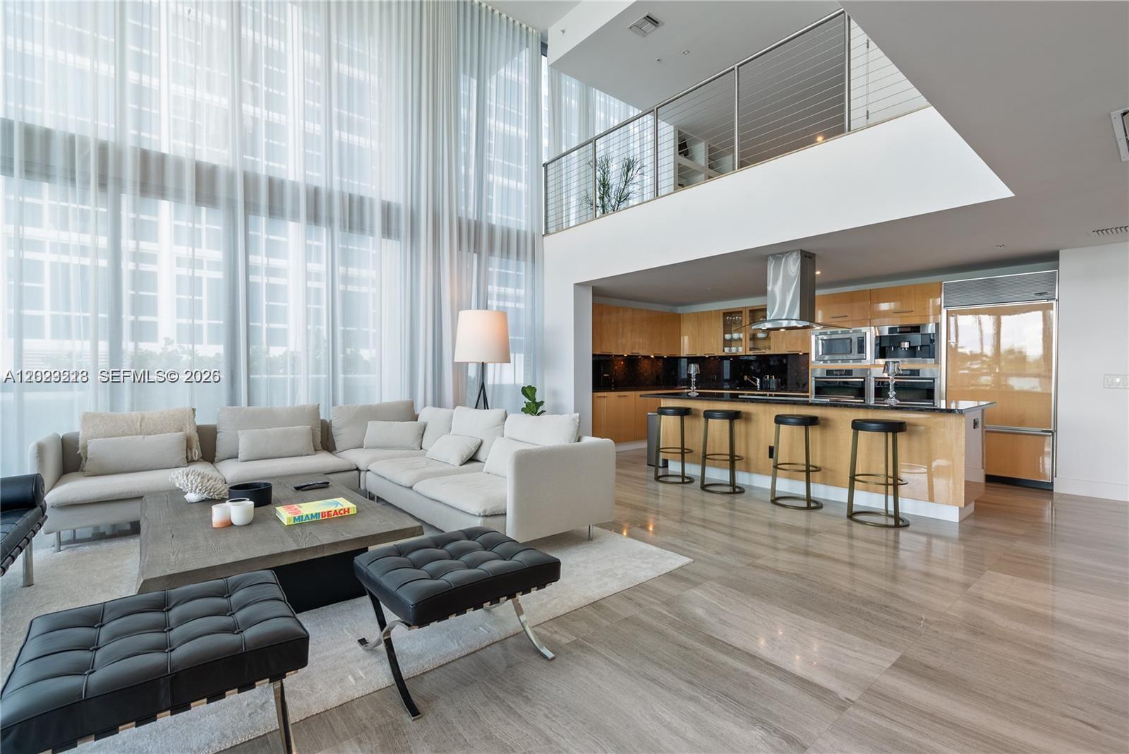 A modern living room and kitchen area feature double-height windows, light wood cabinetry, and tile flooring — 6899 Collins Ave # 202, Miami Beach FL 33141, MIAMI BEACH, FL