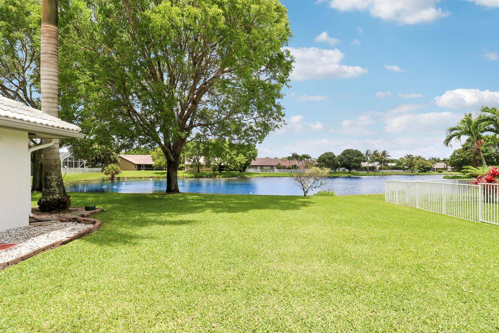 A grassy backyard overlooks a lake with mature trees and neighboring houses in the distance — 7160 Brickyard Circle, Lake Worth FL 33467, LAKE WORTH, FL