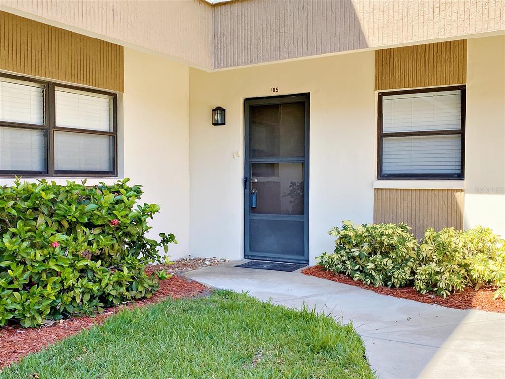 The exterior of a residential unit features a front door numbered 105 and landscaping — 2700 NEBRASKA AVENUE UNIT 5-105, PALM HARBOR, FL