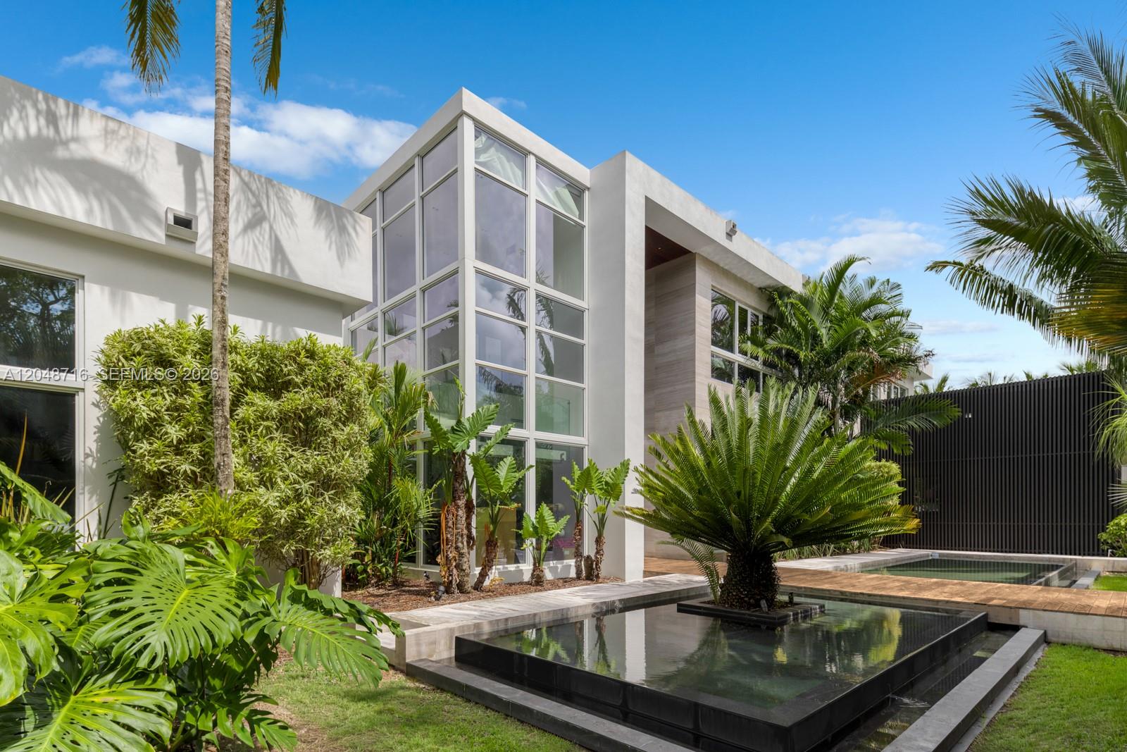A modern white house features a glass-walled corner, a reflecting pool, and lush landscaping — 12900 SW 63rd Ave, Pinecrest FL 33156, PINECREST, FL