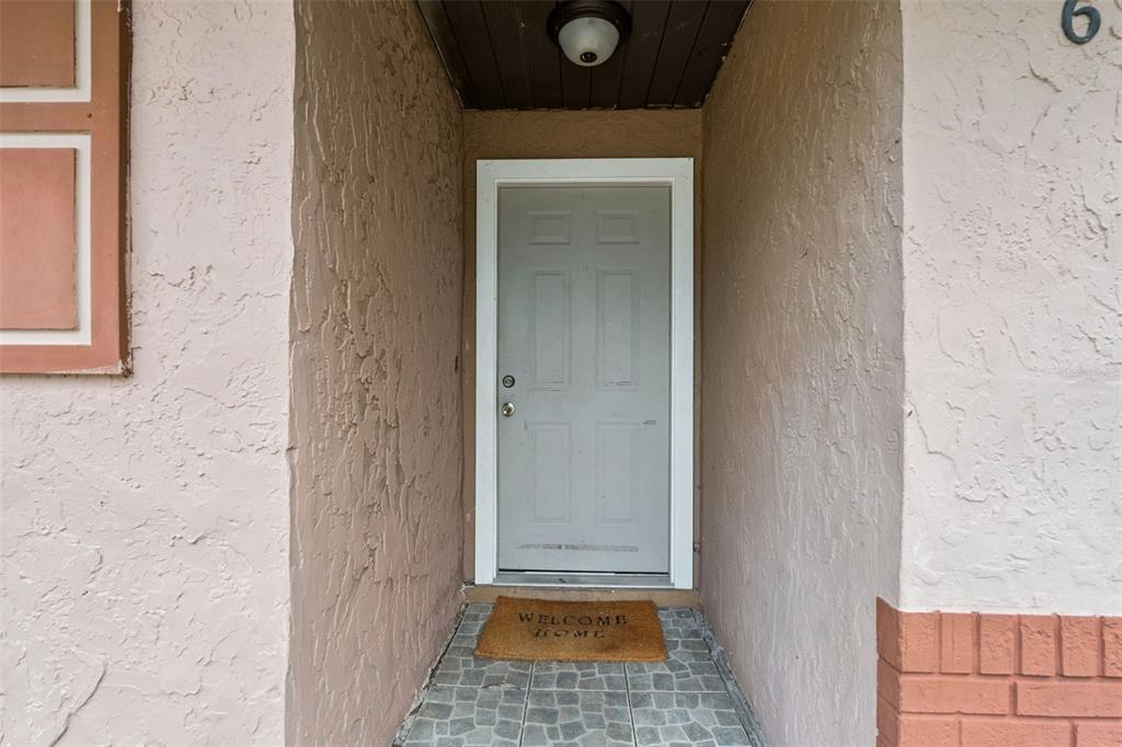 A light gray front door is set within a stucco entryway with a welcome mat — 6835 LONGMEADE LANE, ORLANDO, FL