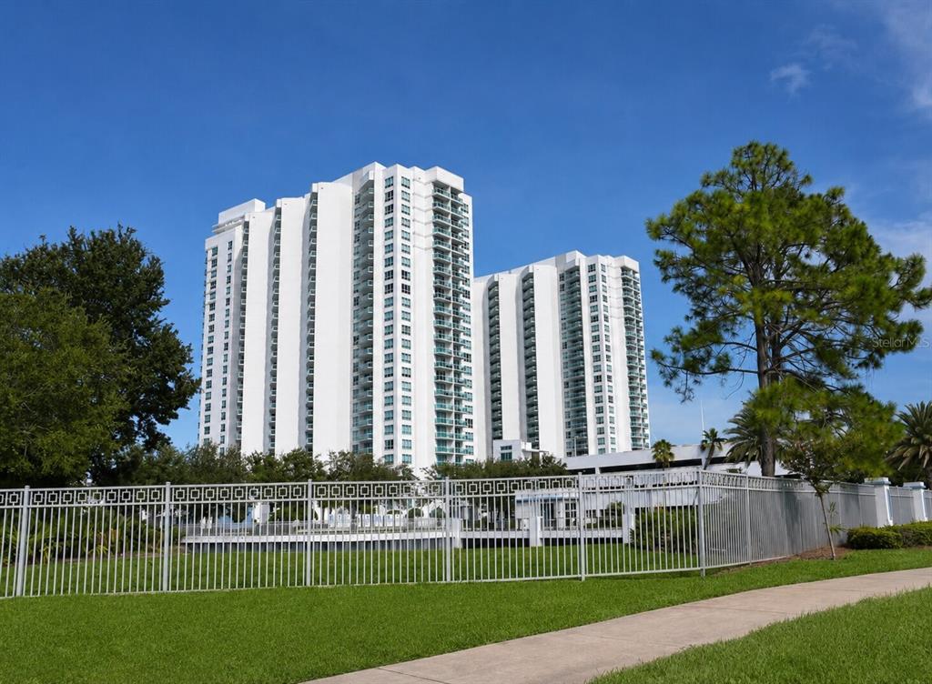 A tall white residential high-rise building stands behind a metal fence and green lawn — 241 RIVERSIDE DRIVE UNIT 2009, HOLLY HILL, FL