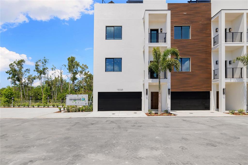 A modern three-story townhouse exterior features white stucco, wood-look siding, and two black garage doors — 10351 2ND STREET N, ST PETERSBURG, FL