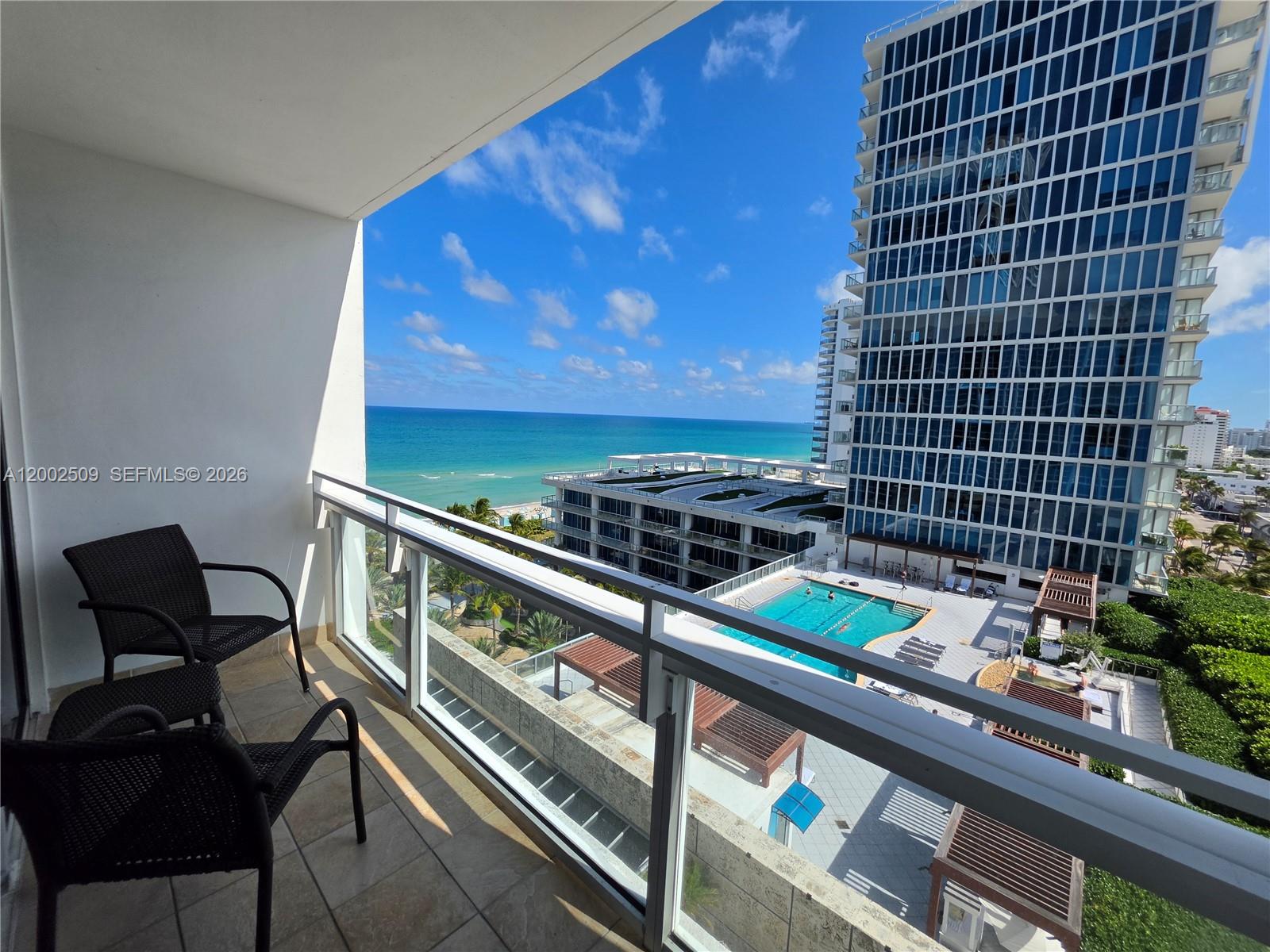 A balcony with two chairs overlooks a swimming pool, a tall glass building, and the ocean — 6801 Collins Ave, Miami Beach FL 33141, MIAMI BEACH, FL