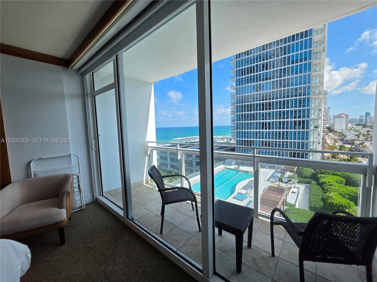 A room with a beige armchair opens onto a balcony overlooking a pool and ocean — 6801 Collins Ave, Miami Beach FL 33141, MIAMI BEACH, FL