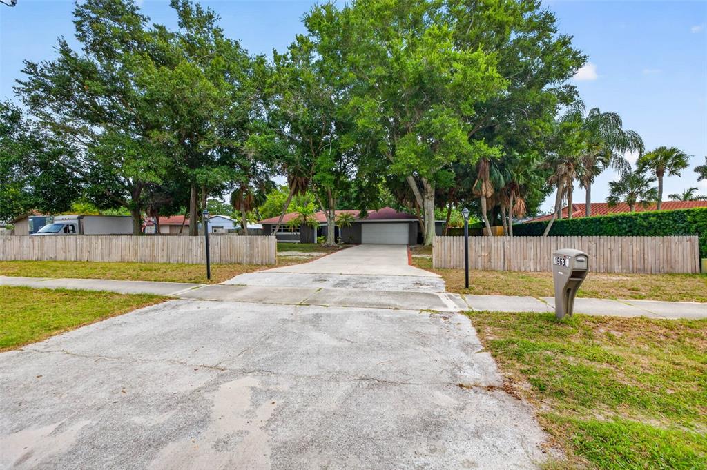 A single-story house with a concrete driveway and a wooden fence sits behind large trees — 1963 BELLEAIR ROAD, CLEARWATER, FL