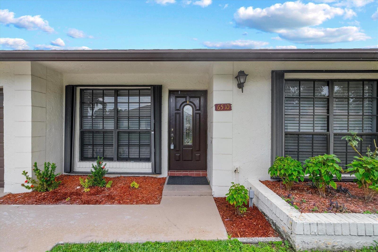 The exterior of a house features a dark front door, two windows, and landscaping — 6510 Olivewood Circle, FL 33463, FL