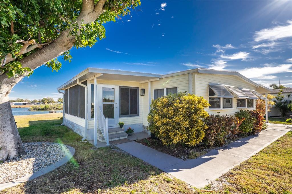 A light-yellow mobile home with a screened-in porch sits beside a lake under a blue sky — 1300 SANDERLING DRIVE, ENGLEWOOD, FL