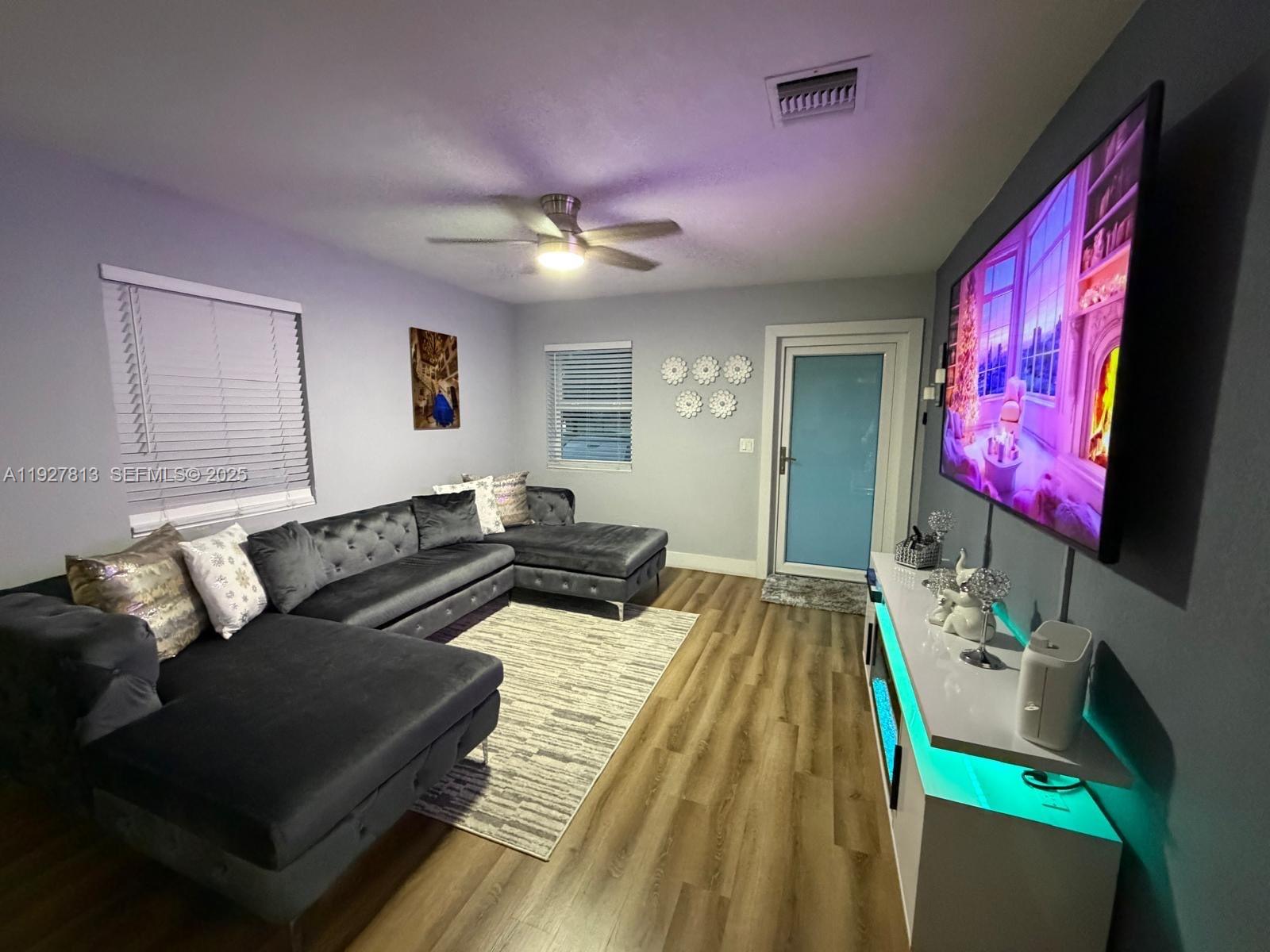A living room features a grey sectional sofa, wood-look flooring, and a wall-mounted television — 100 NE 171st Ter, Miami FL 33162, MIAMI, FL