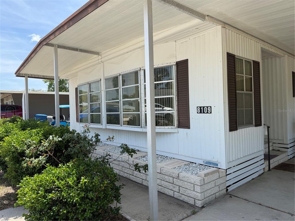 The exterior of a white mobile home features brown shutters, a covered porch, and landscaping — 8109 LITTLE TEE LANE, BROOKSVILLE, FL