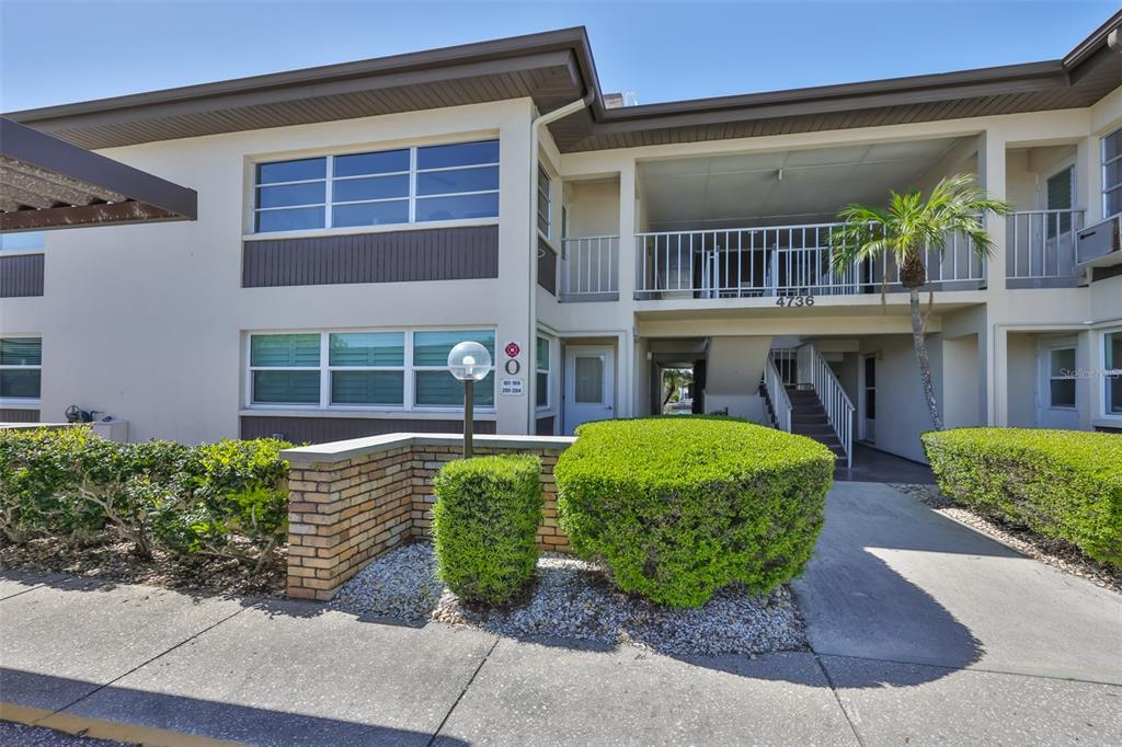 The exterior of a two-story apartment building features a concrete walkway, landscaping, and stairs — 4736 JASPER DR UNIT 103, NEW PORT RICHEY, FL