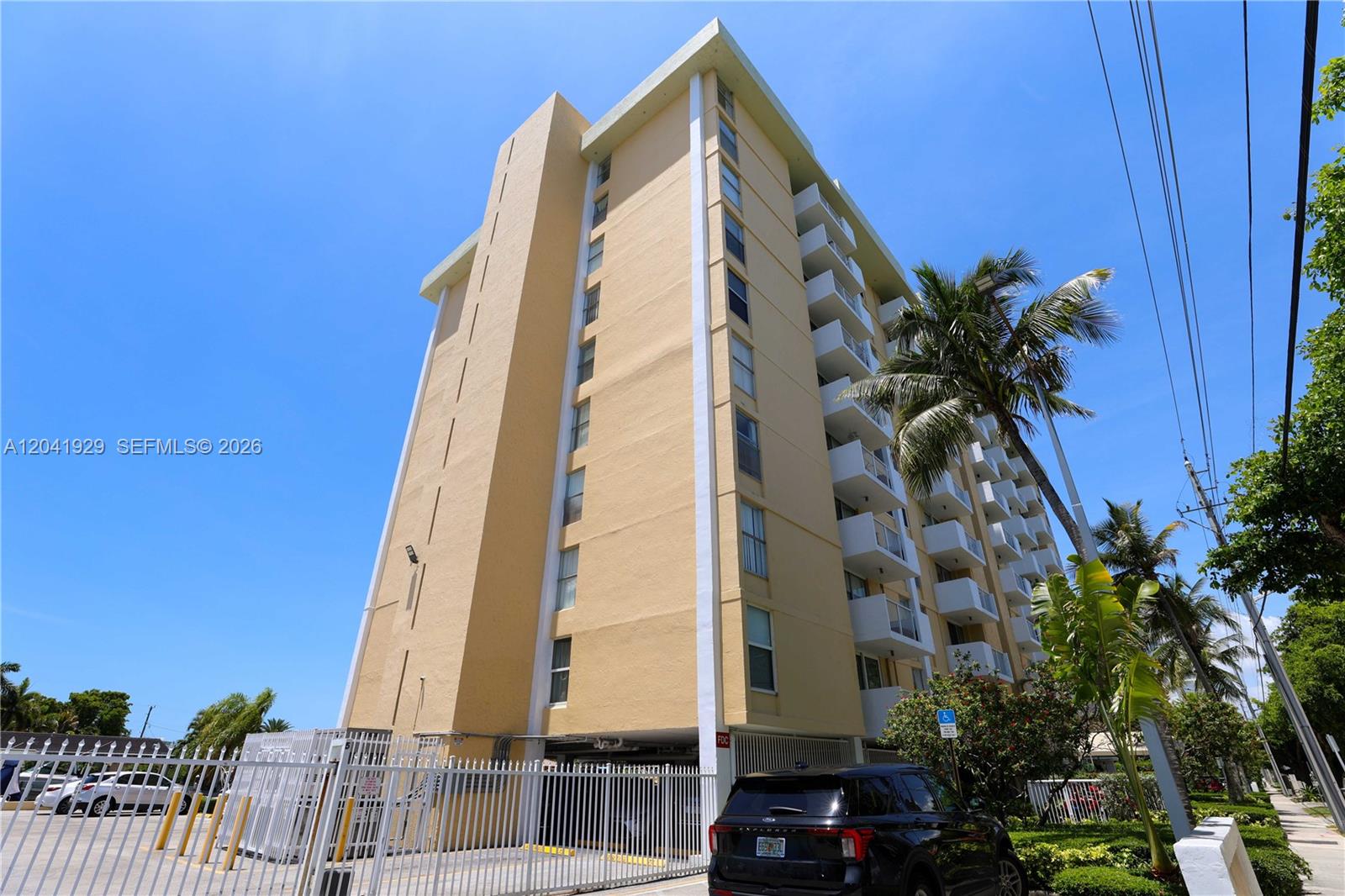 A low-angle exterior view of a multi-story tan apartment building with balconies and a parking area — 2000 NE 135th St # 906, North Miami FL 33181, NORTH MIAMI, FL