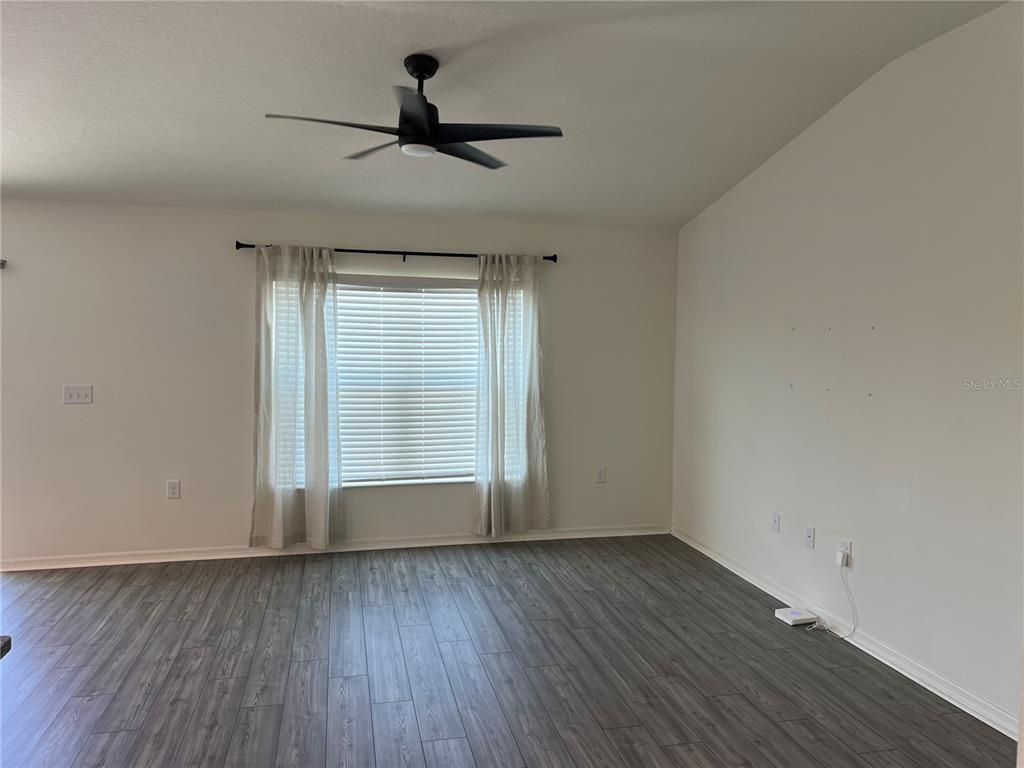 An empty living room features gray wood-look flooring, white walls, a ceiling fan, and window curtains — 3838 WHITETAIL COURT, ST CLOUD, FL