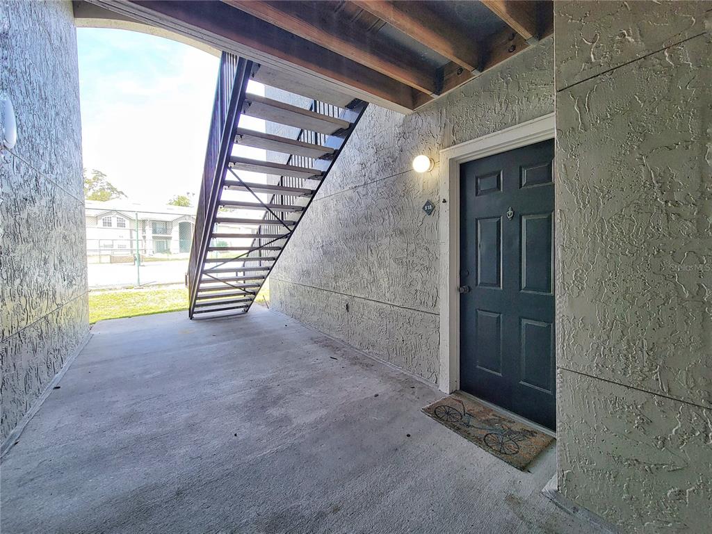 A dark door with a welcome mat sits under an outdoor staircase on a concrete patio — 3705 SW 27TH STREET UNIT 818, GAINESVILLE, FL