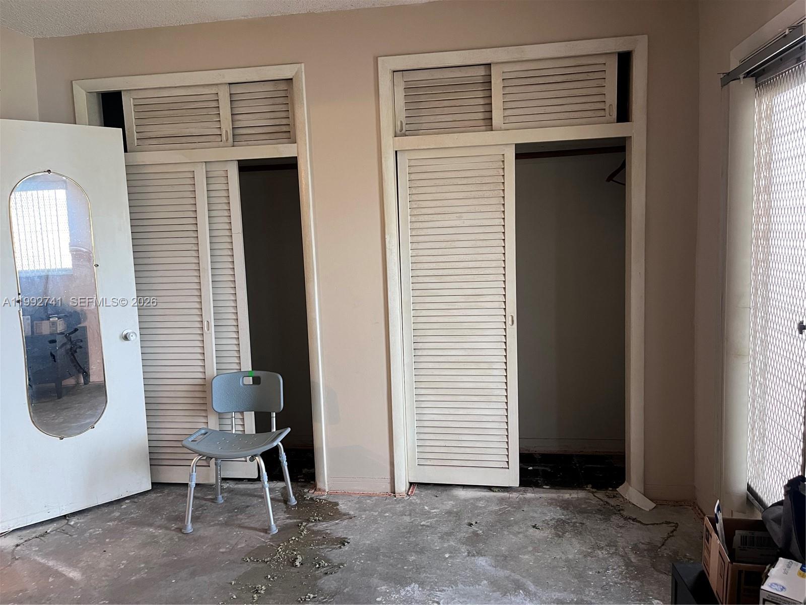 A room with two louvered-door closets, a gray shower chair, and an unfinished concrete floor — 1012 SW 11th St # 9N, Hallandale Beach FL 33009, HALLANDALE BEACH, FL
