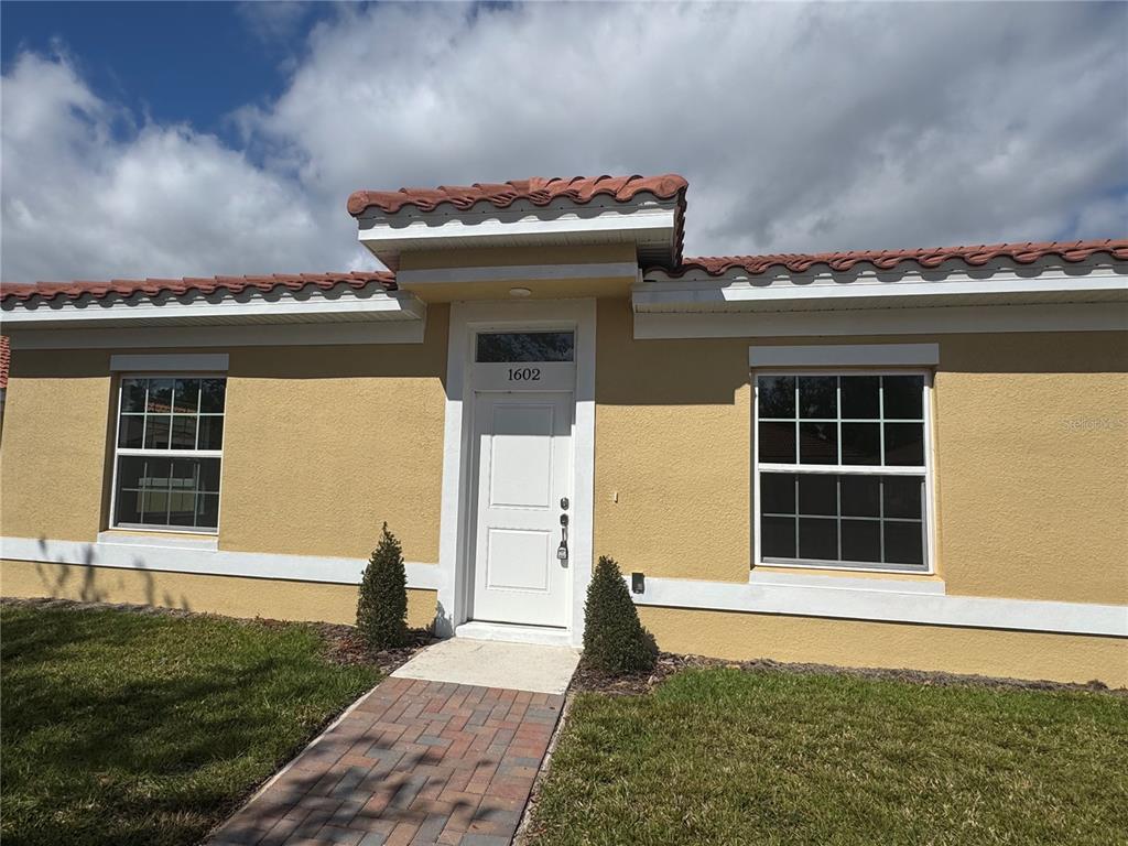 A yellow stucco house exterior features a white front door, two windows, and tile roofing — 1602 CUMIN DRIVE UNIT 1602, POINCIANA, FL