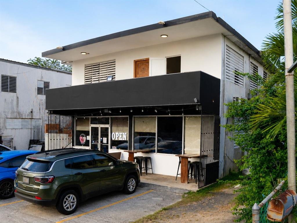 A two-story commercial building with a black awning, outdoor seating, and a parked green SUV — #2 St. CARR. JUAQUIN “YIYE” AVILA, QUEBRADILLAS, PR