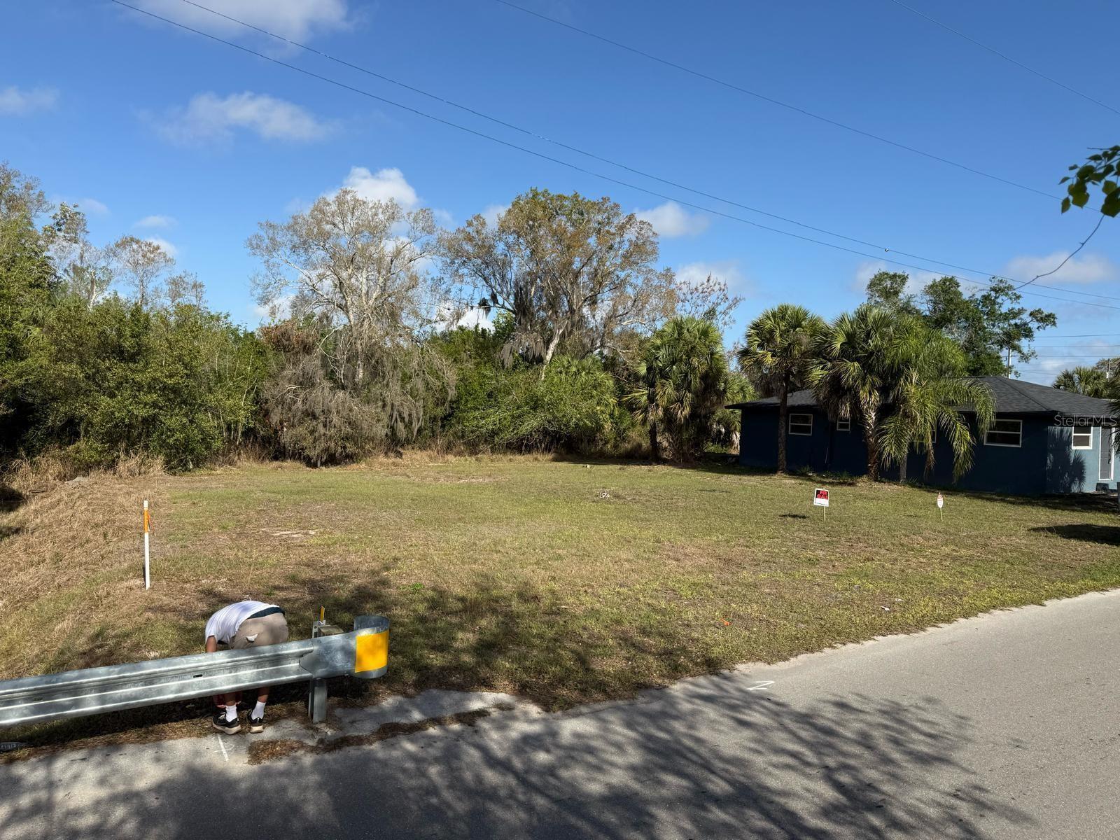 A person stands near a guardrail in front of a vacant, grassy residential lot — Xxxx Singleton Avenue, FL 34266, FL
