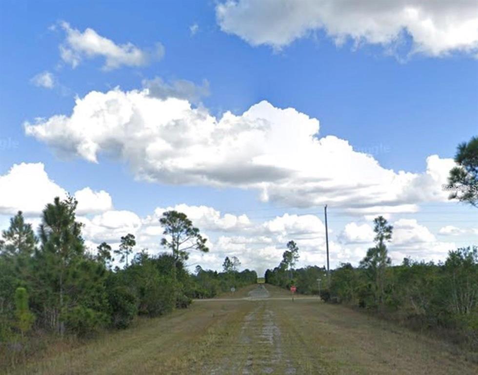A dirt road leads toward an intersection surrounded by trees under a cloudy blue sky — 1008 CALLIANDRA DRIVE, INDIAN LAKE ESTATES, FL