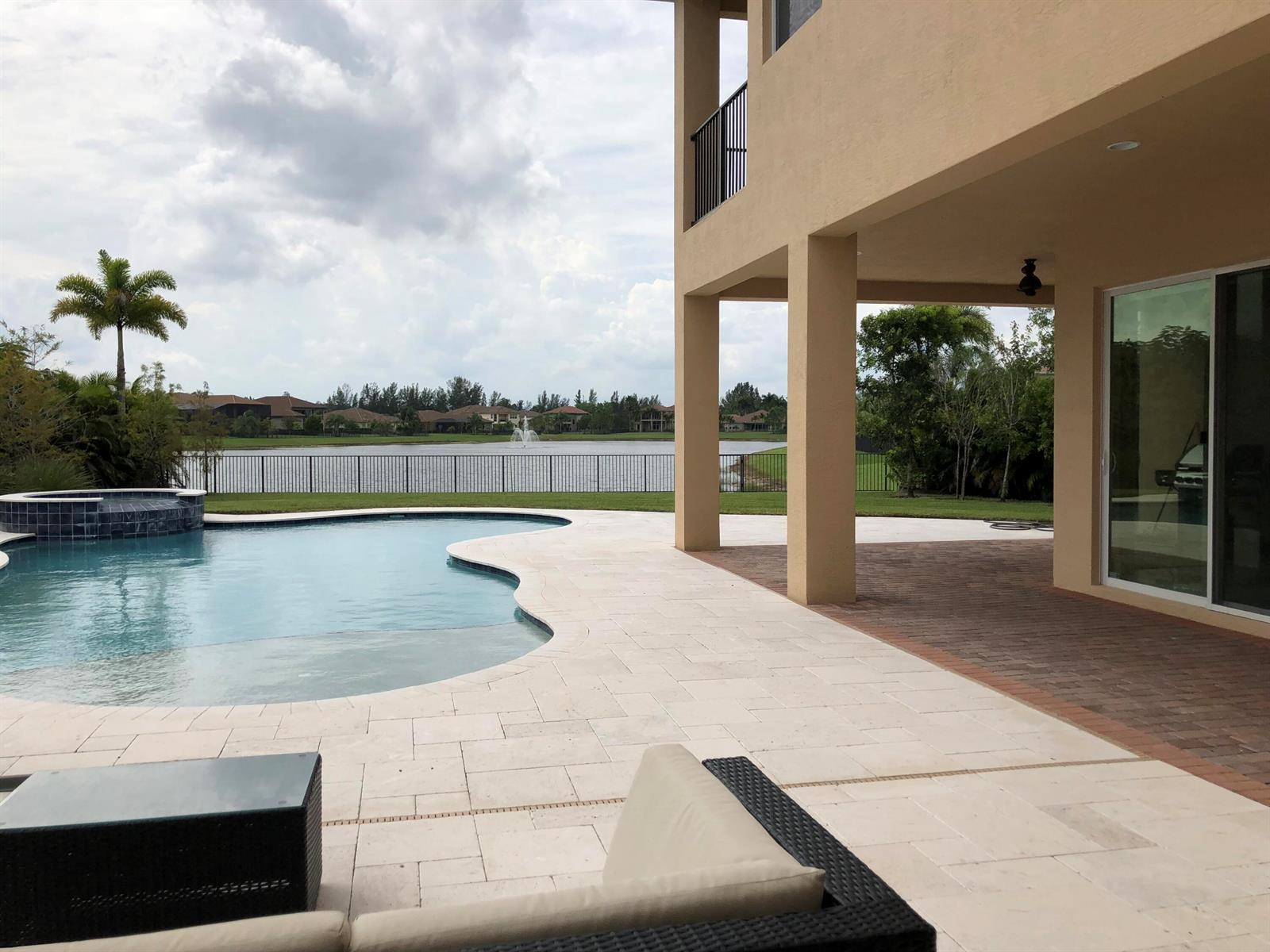 A swimming pool and patio overlook a lake behind a two-story house with a balcony — 7793 Arbor Crest Way, West Palm Beach FL 33412, WEST PALM BEACH, FL