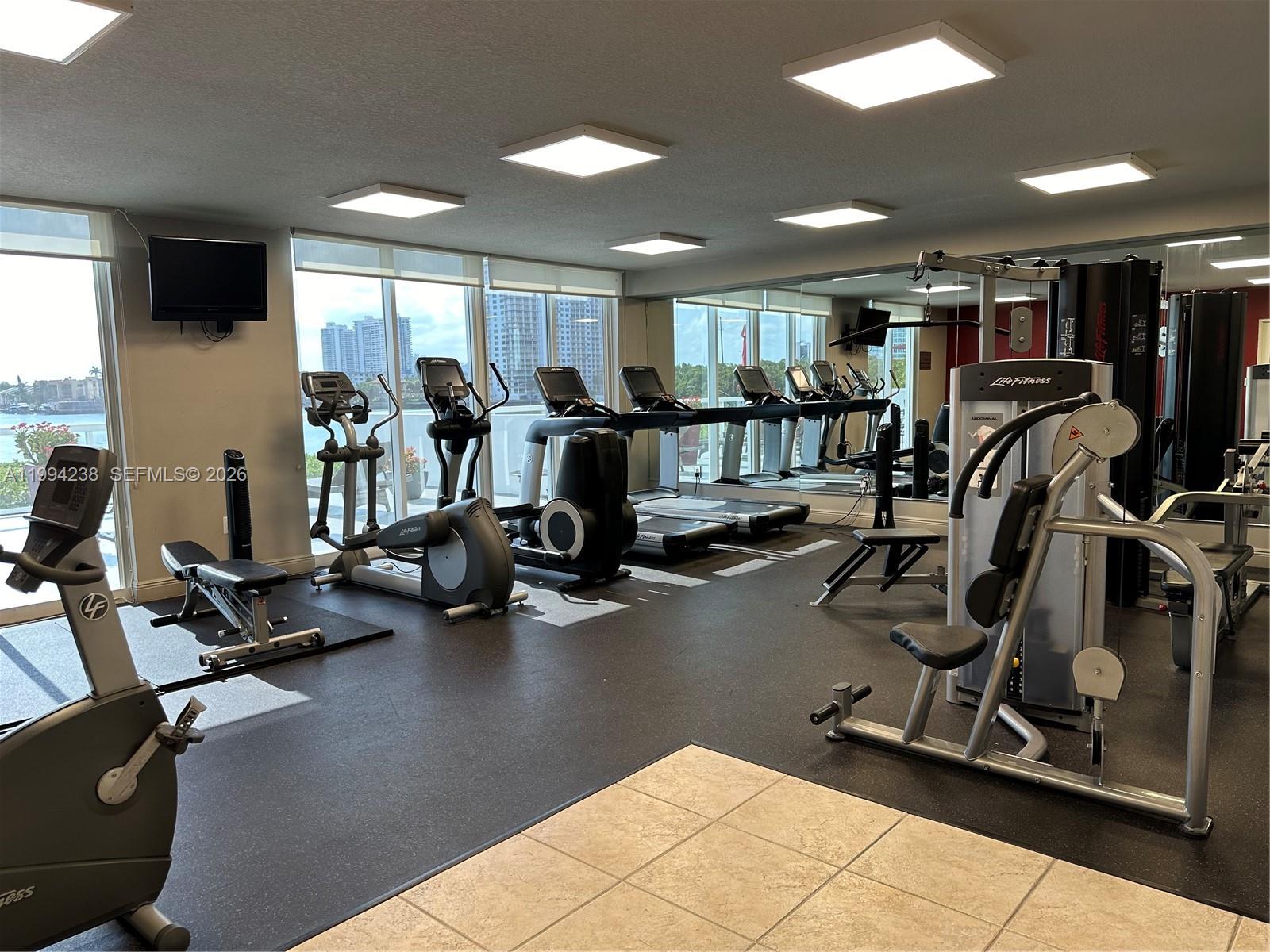 A gym features various exercise machines, large windows with city views, and a mirrored wall — SUNNY ISLES BEACH, FL