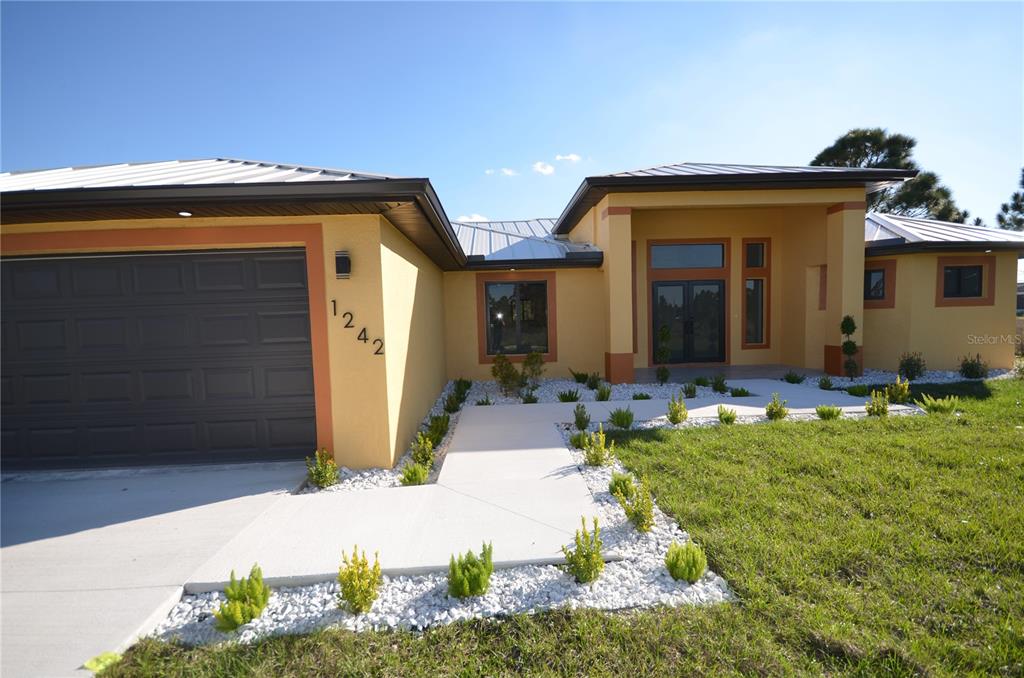 A single-story house with a yellow stucco exterior, metal roof, and a two-car garage — 1242 HATHOR STREET E, LEHIGH ACRES, FL