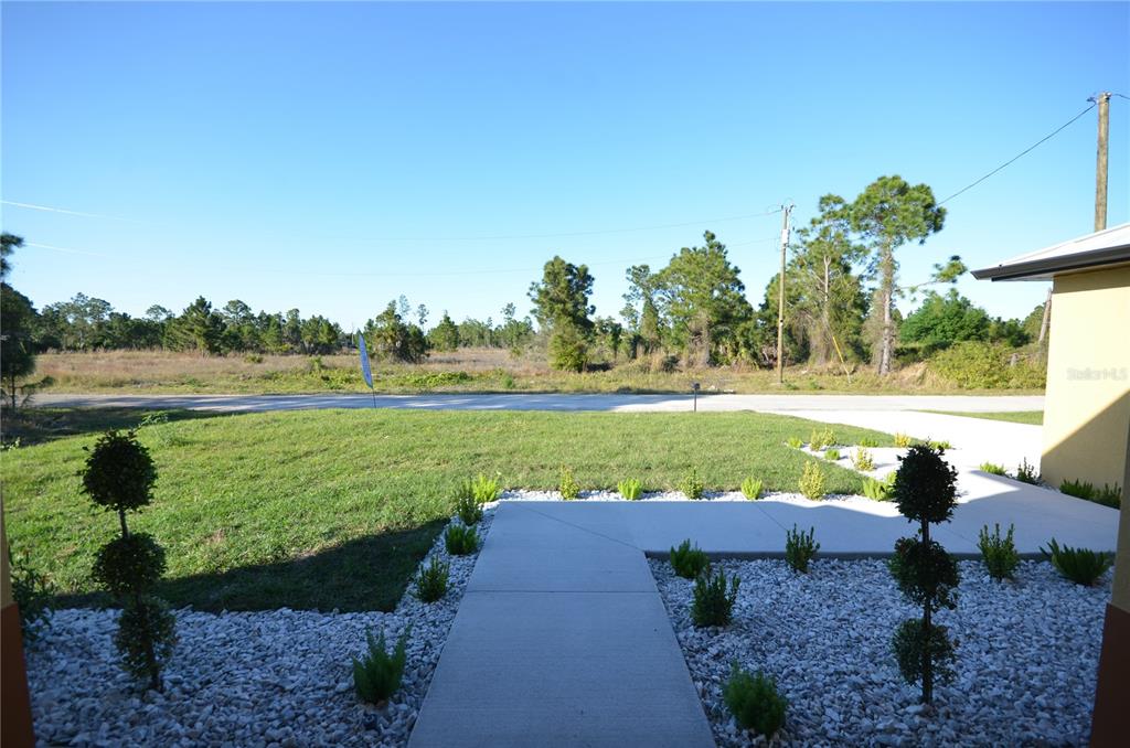 A concrete walkway leads through a landscaped yard toward an open, grassy field — 1242 HATHOR STREET E, LEHIGH ACRES, FL