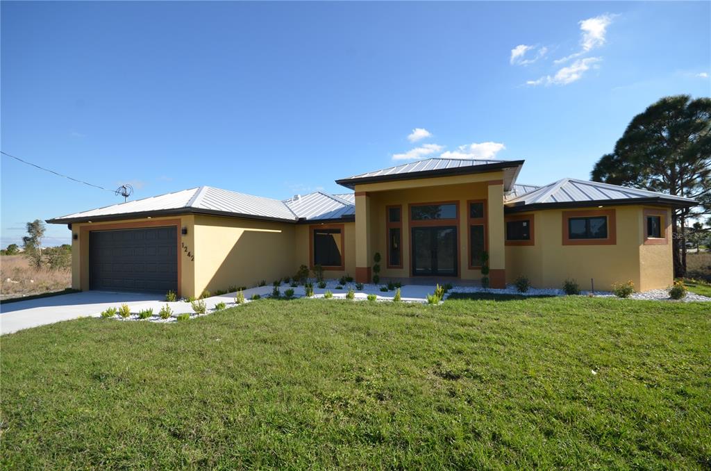 A single-story house with a yellow stucco exterior, metal roof, and attached two-car garage — 1242 HATHOR STREET E, LEHIGH ACRES, FL
