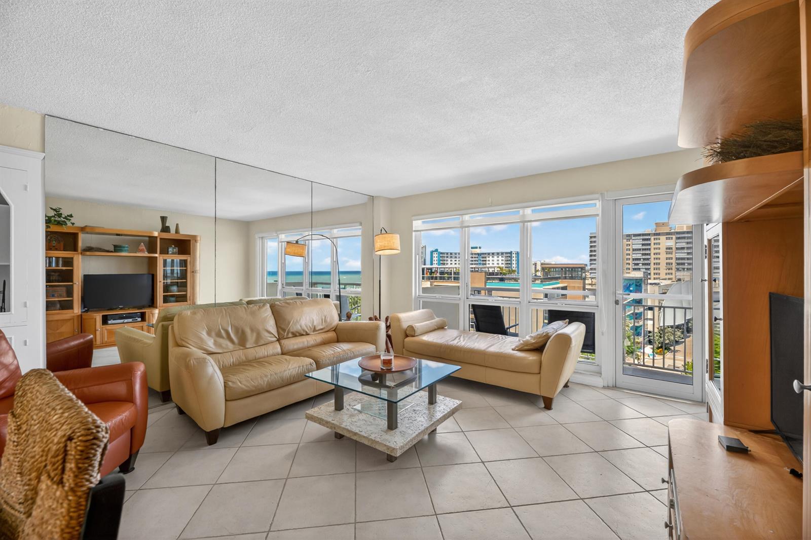 A living room with beige leather furniture, a glass coffee table, mirrored walls, and balcony views — 133 N Pompano Beach Boulevard # 708, Pompano Beach FL 33062, POMPANO BEACH, FL