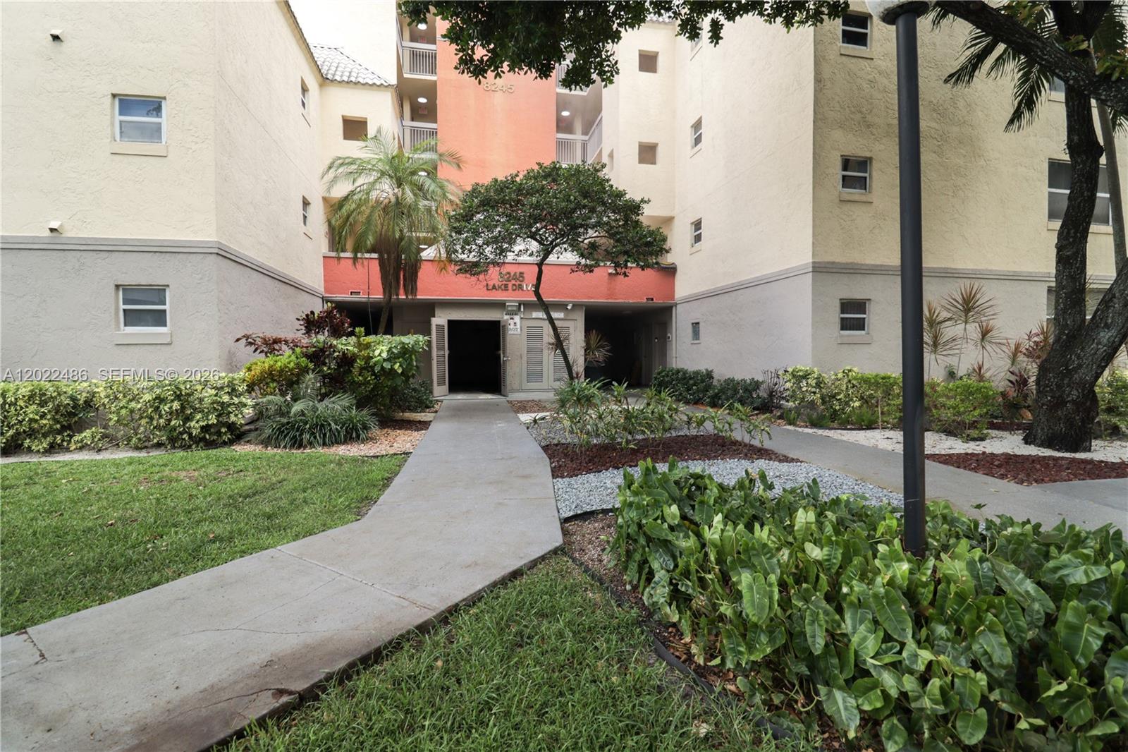 A concrete walkway leads to the entrance of a multi-story apartment building with landscaping — 8245 Lake Dr # 308, Doral FL 33166, DORAL, FL