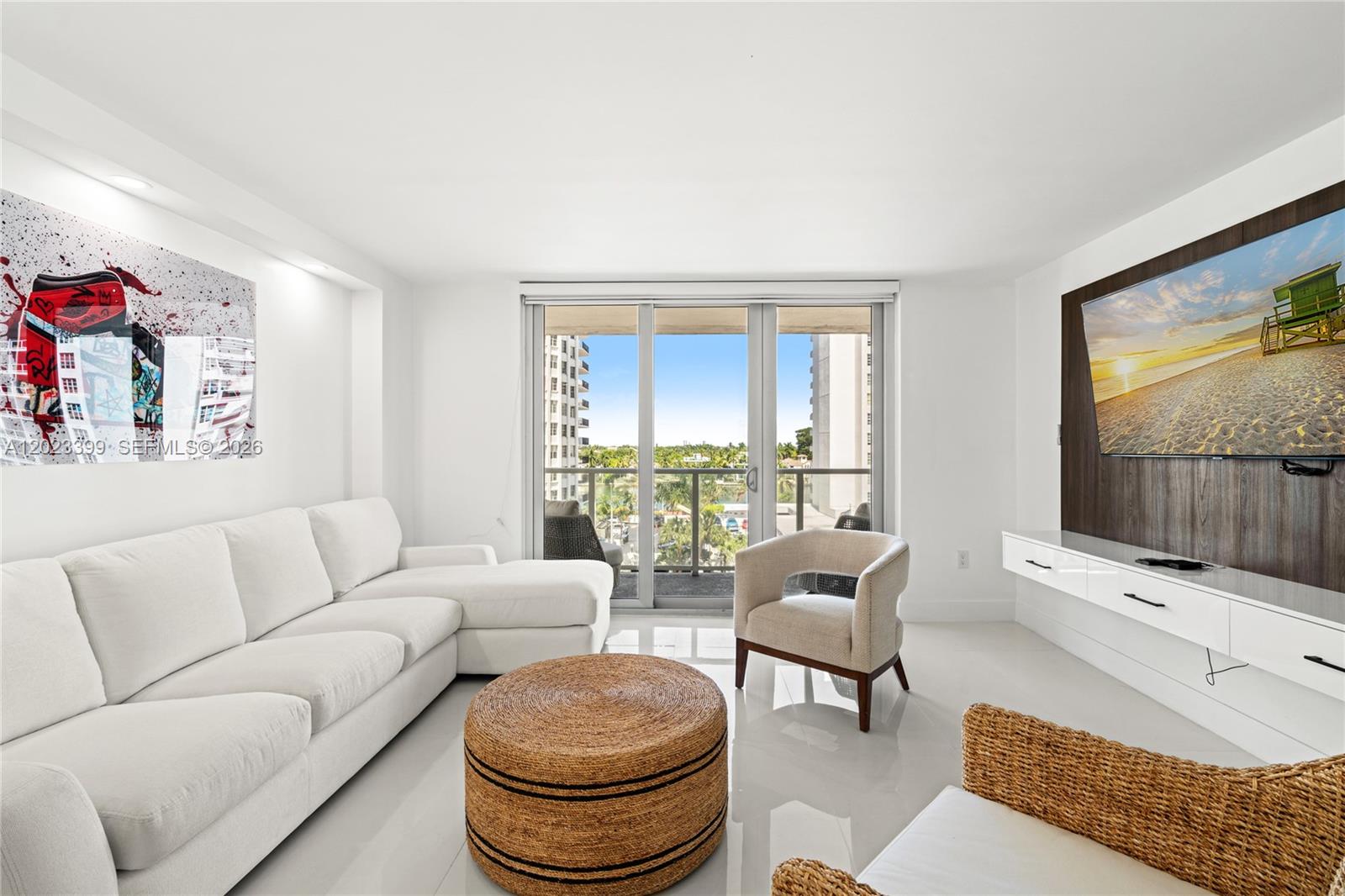 A bright living room features a white sectional sofa, a round woven ottoman, and a balcony — 5701 Collins Ave # 503, Miami Beach FL 33140, MIAMI BEACH, FL