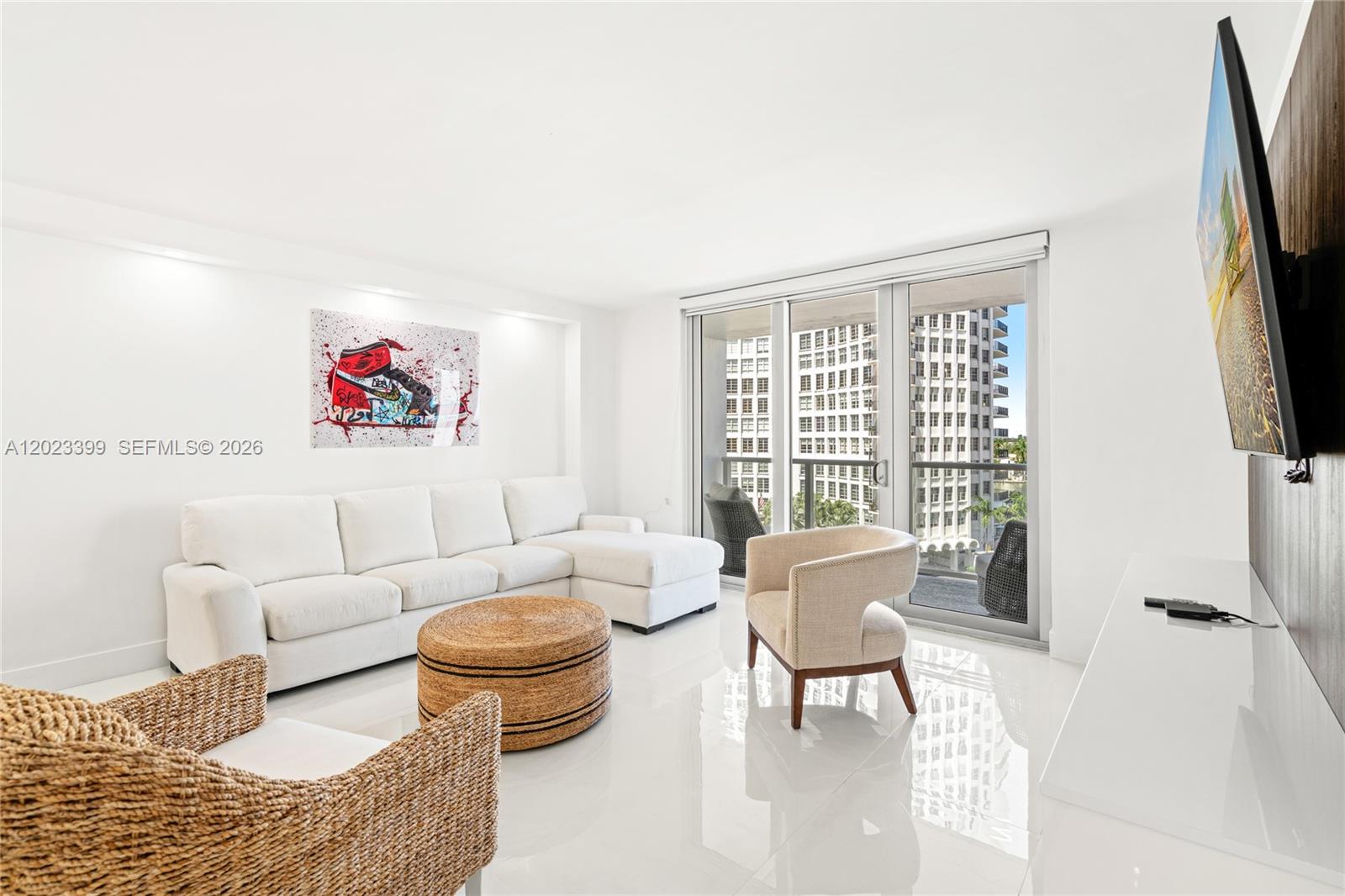 A bright living room features a white sectional sofa, woven furniture, and a balcony view — 5701 Collins Ave # 503, Miami Beach FL 33140, MIAMI BEACH, FL