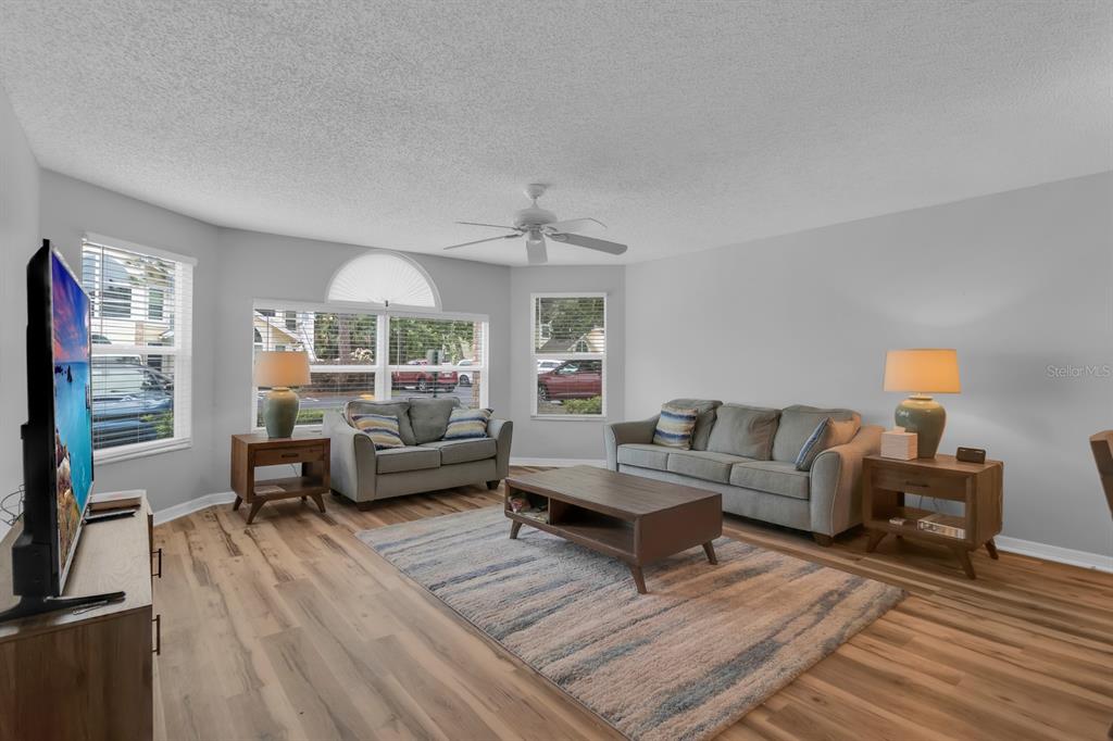 A living room features light wood-look flooring, a gray sofa and loveseat, and large windows — 5004 LAGUNA BAY CIRCLE UNIT 49, KISSIMMEE, FL