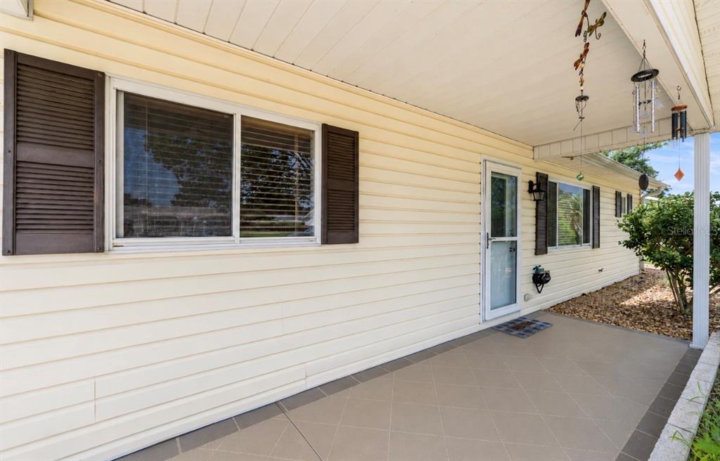 A covered porch with light-colored siding, dark shutters, and tiled flooring leads to a front door — 10358 SE 178 PLACE, SUMMERFIELD, FL