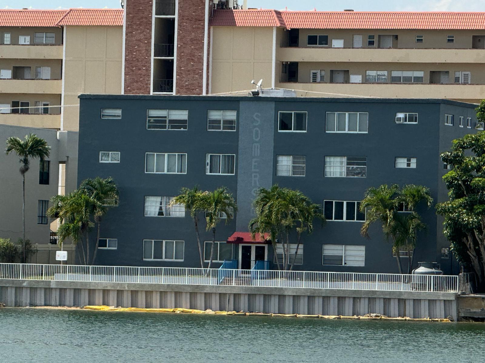 A gray four-story apartment building sits along a waterfront with a white metal fence — 1060 Crystal Lake Drive # 101, Deerfield Beach FL 33064, DEERFIELD BEACH, FL