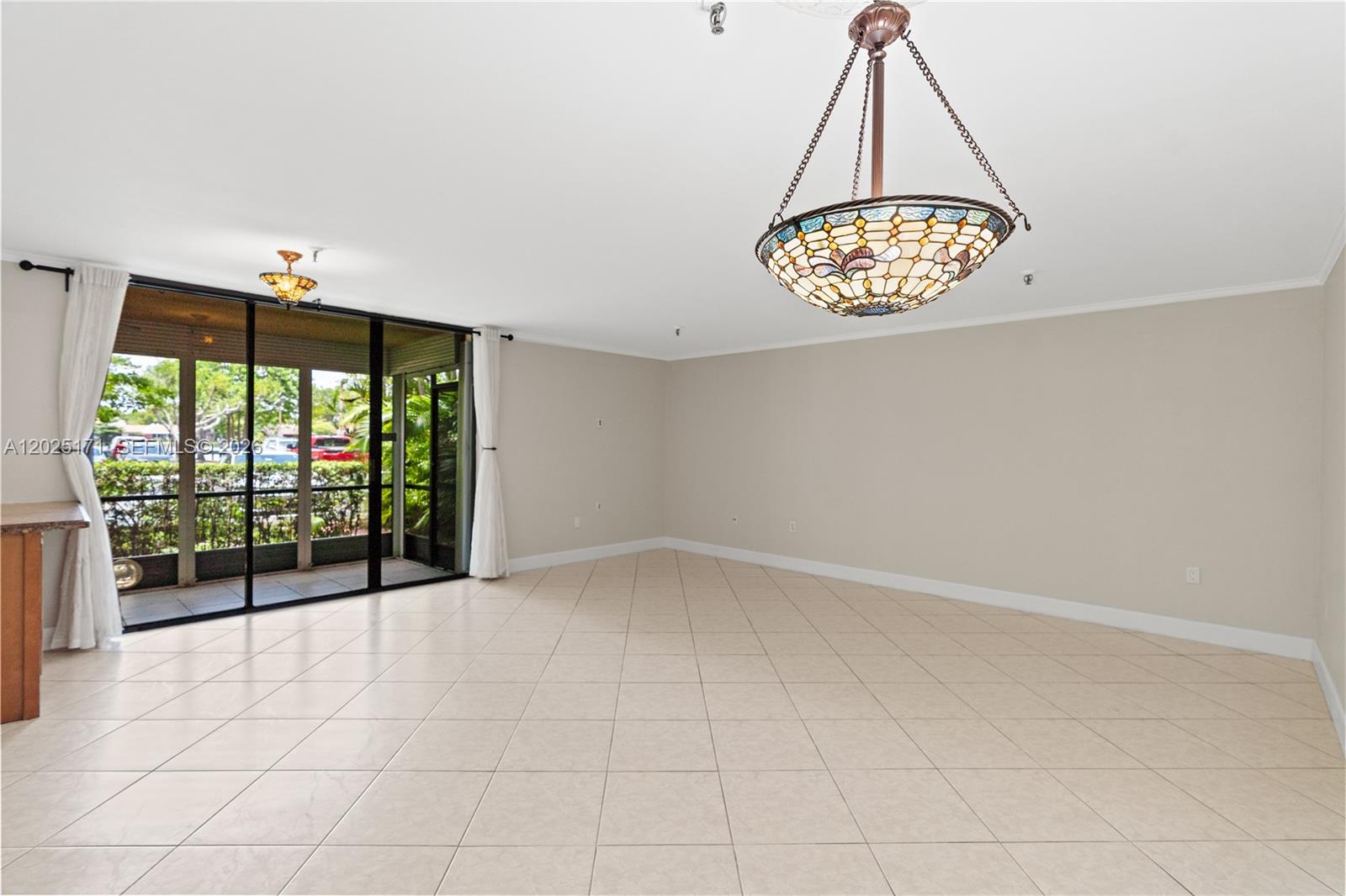 An empty room features beige tile flooring, a stained-glass chandelier, and sliding glass doors — 671 NE 195th St # 121E, Miami FL 33179, MIAMI, FL